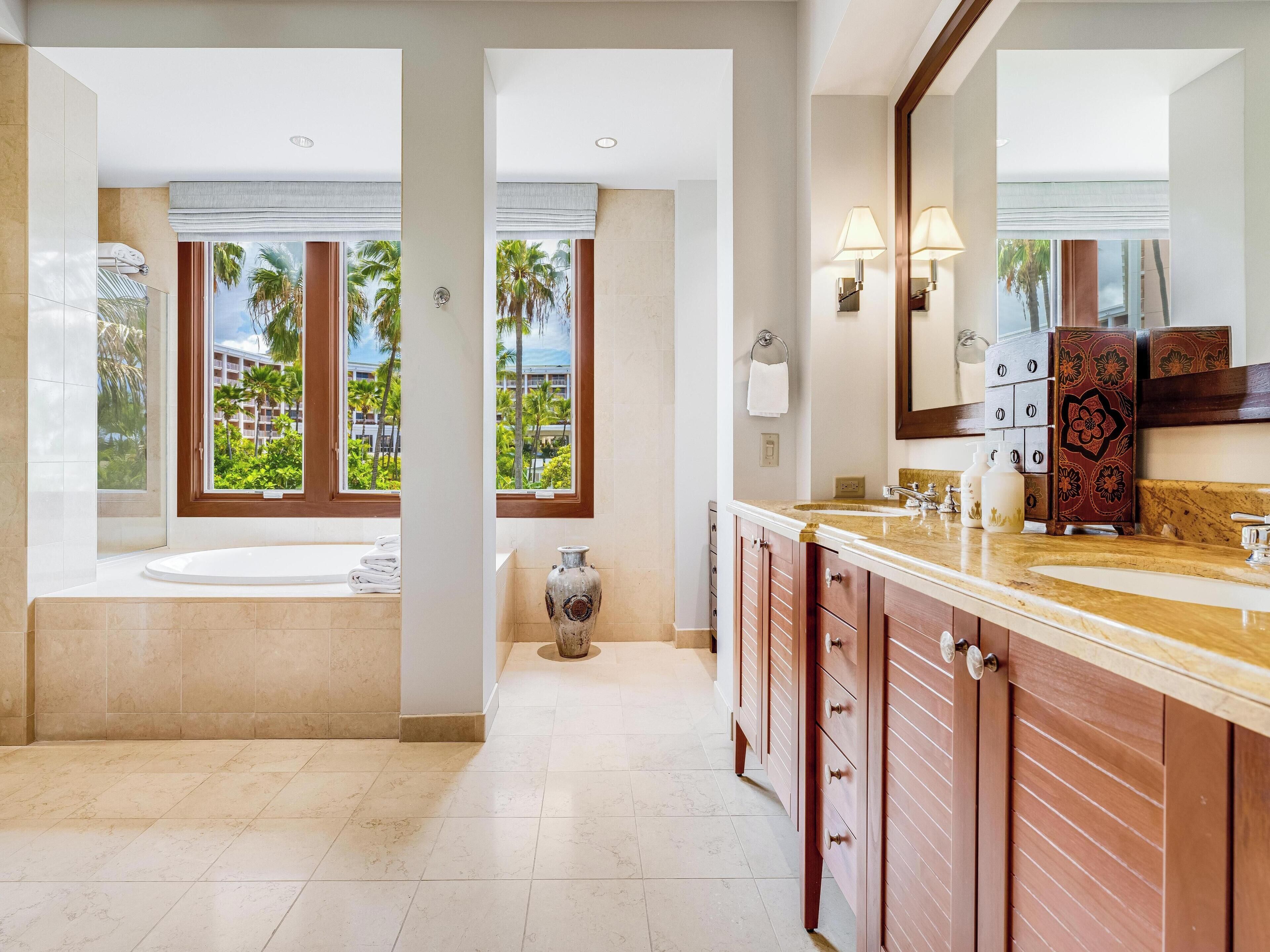 A modern bathroom with a large bathtub, a vanity with a mirror, and a view of palm trees and a building through the windows.