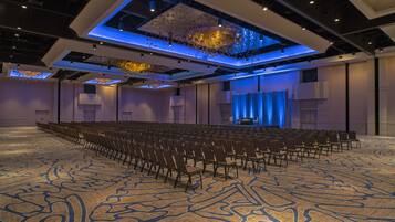 A large conference room with rows of chairs, a patterned carpet, and a stage with a blue curtain.