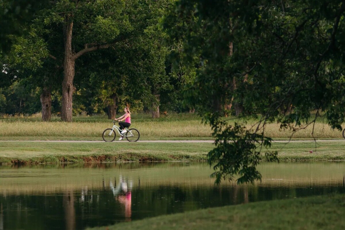 Two cyclists on a path beside a calm body of water, surrounded by trees and grassy areas.