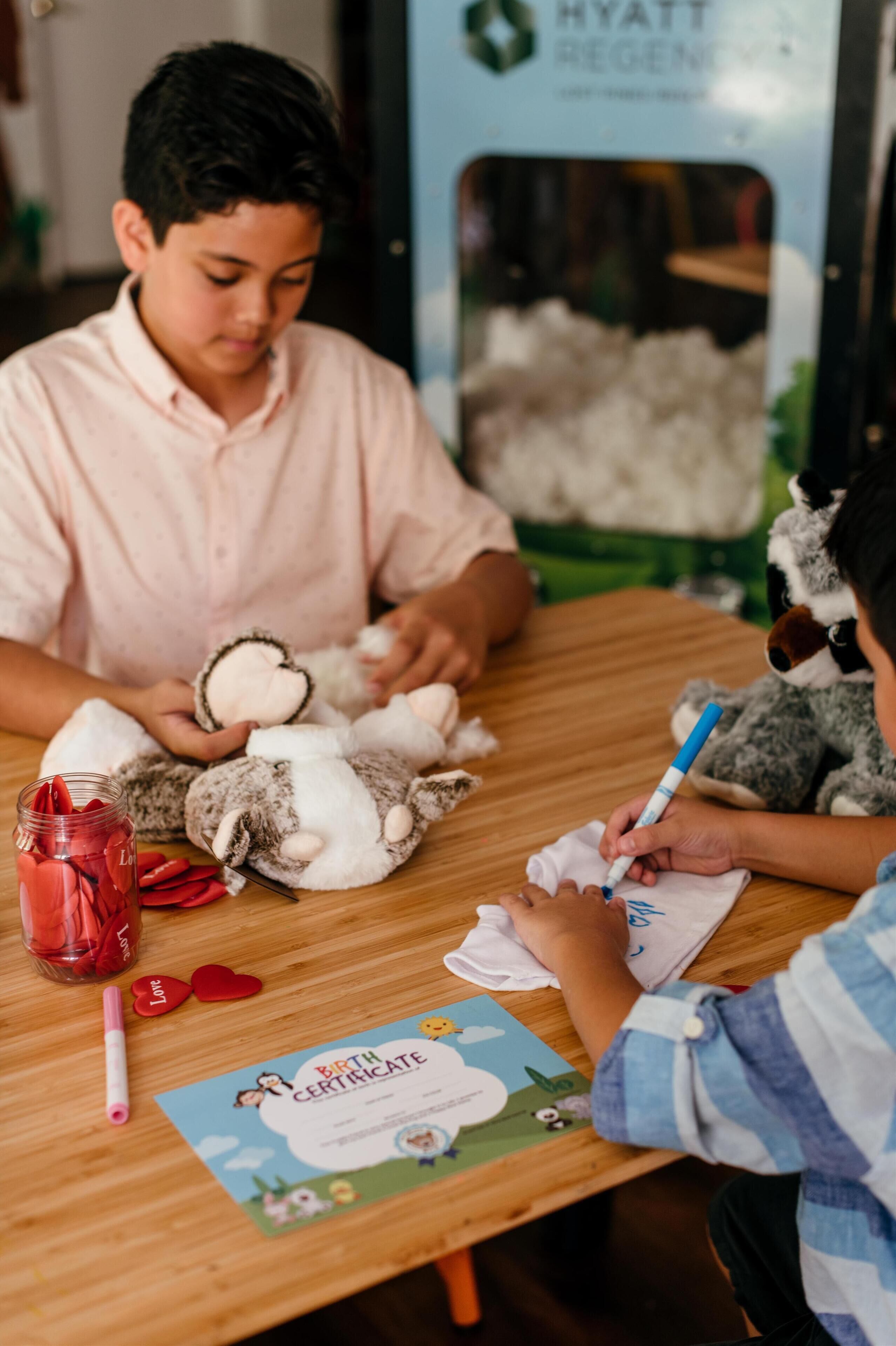 A child drawing on a birth certificate at a wooden table with stuffed animals and a jar of strawberries.