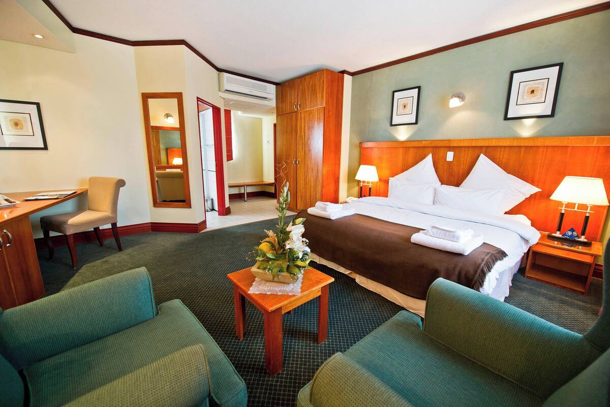 Quarto de hotel com uma cama, duas poltronas verdes, uma pequena mesa de madeira com um vaso, uma escrivaninha com cadeira e um guarda-roupa.