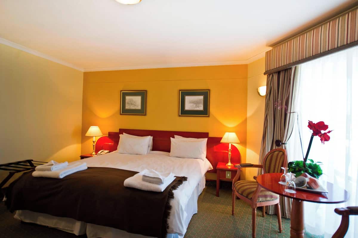 Quarto de hotel com uma cama grande, duas luminárias de cabeceira, uma mesinha com uma planta e uma cadeira.