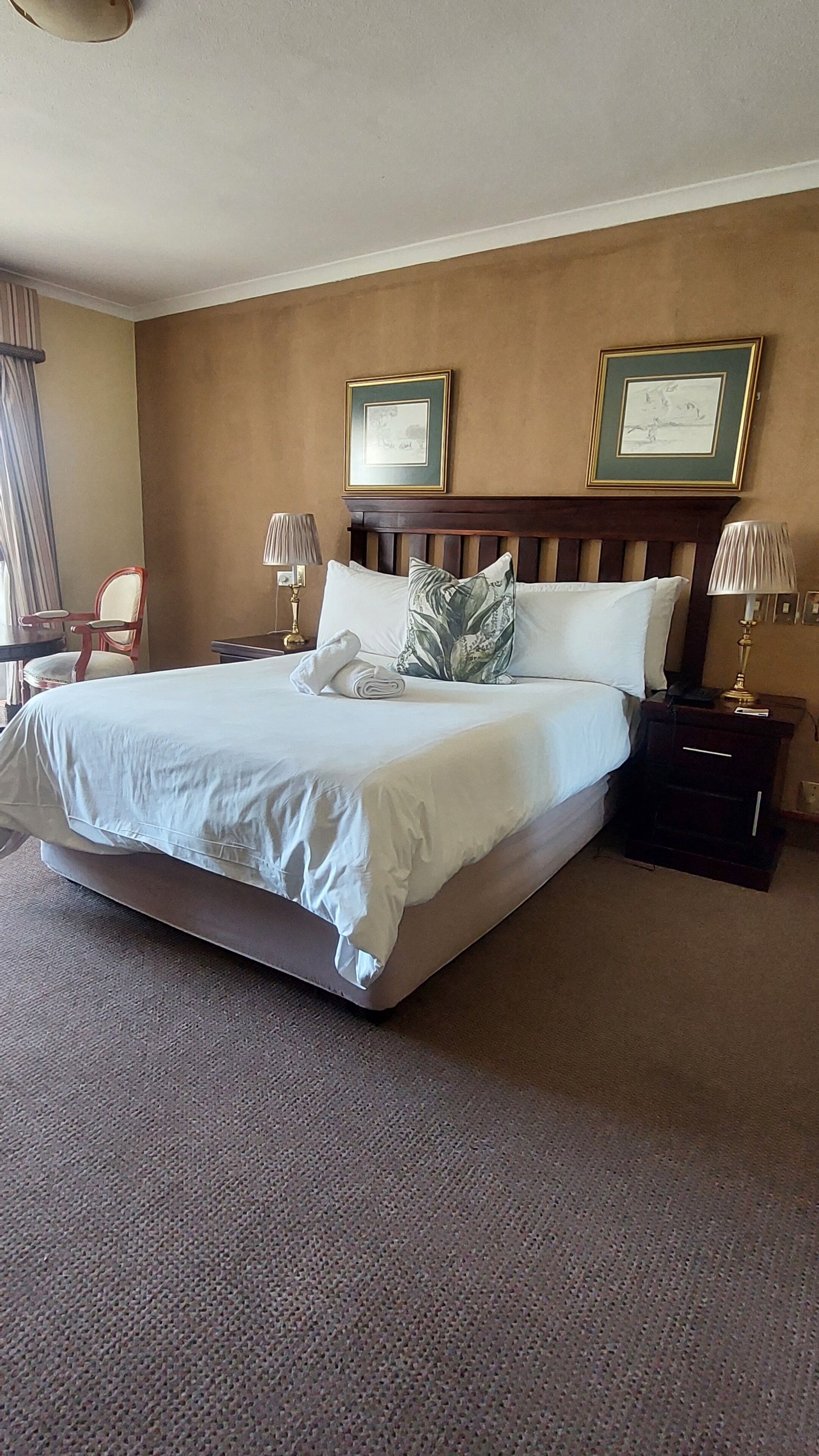 A hotel room with a large bed, two framed pictures on the wall, a nightstand with a lamp, and a chair.