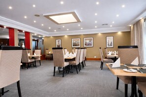 Een restaurantgedeelte in een hotel met meerdere tafels en stoelen, plafondlampen en ingelijste foto's aan de muur.
