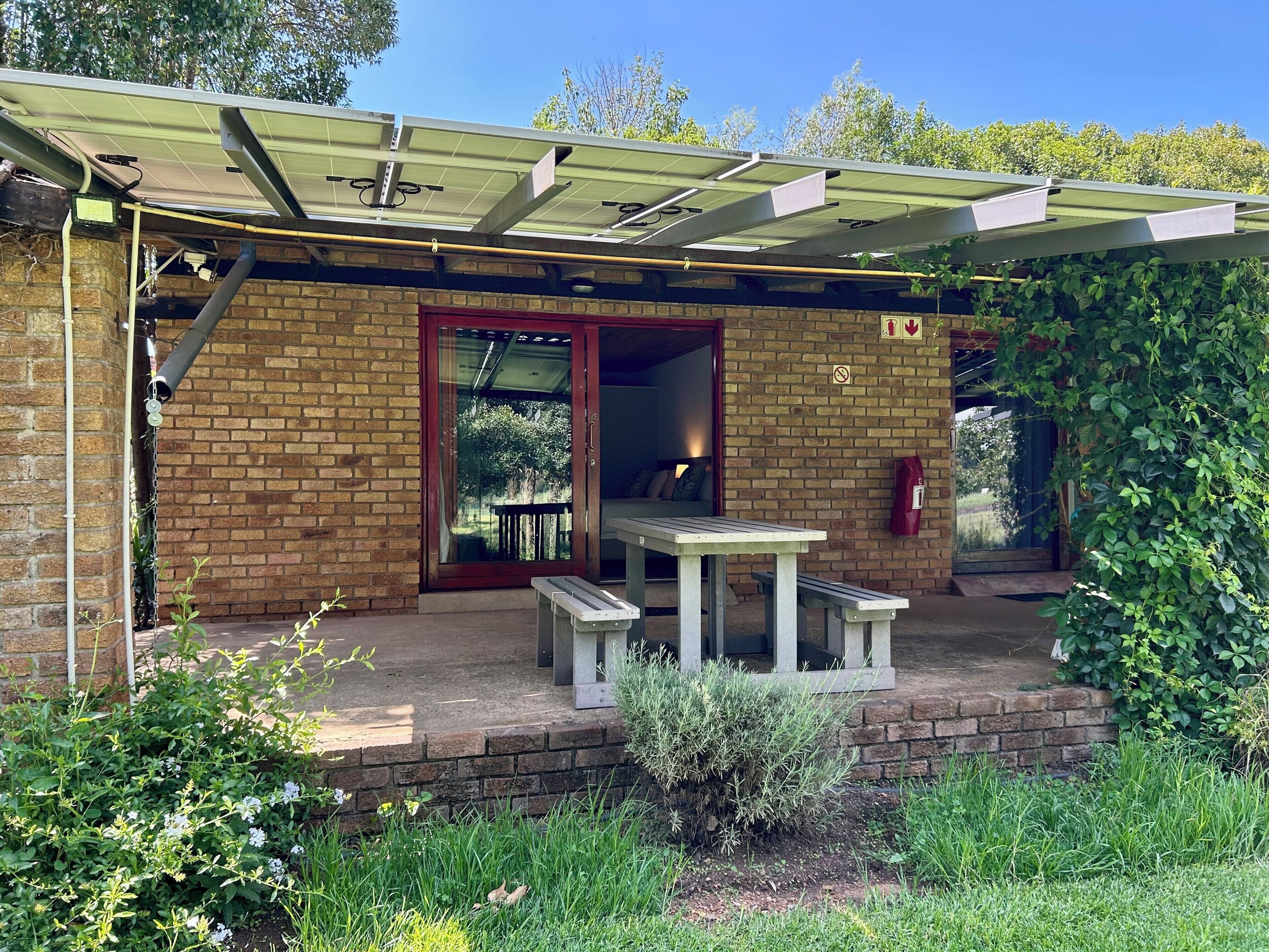 A brick building with a glass door, a covered patio with a table and benches, and a garden area.