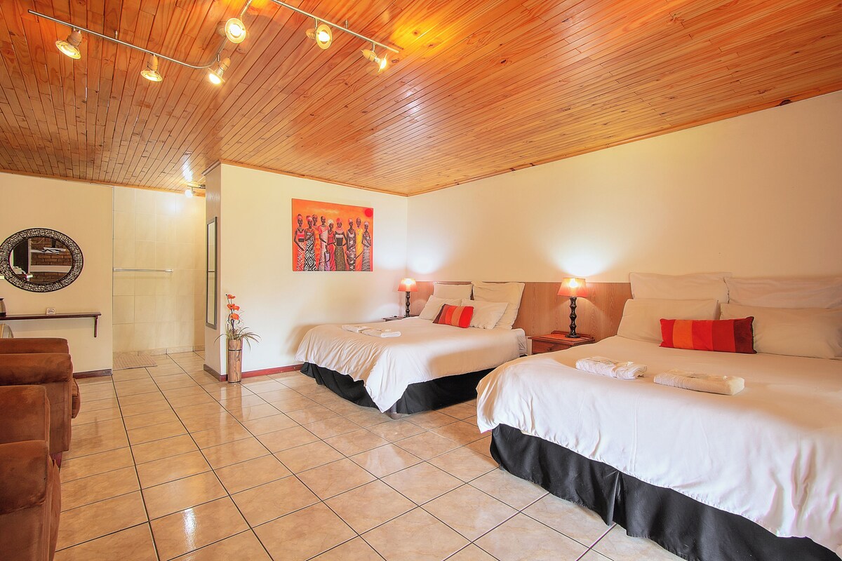 Habitación de hotel con dos camas, techo de madera, suelo de baldosas, un cuadro en la pared y una mesita con una lámpara.