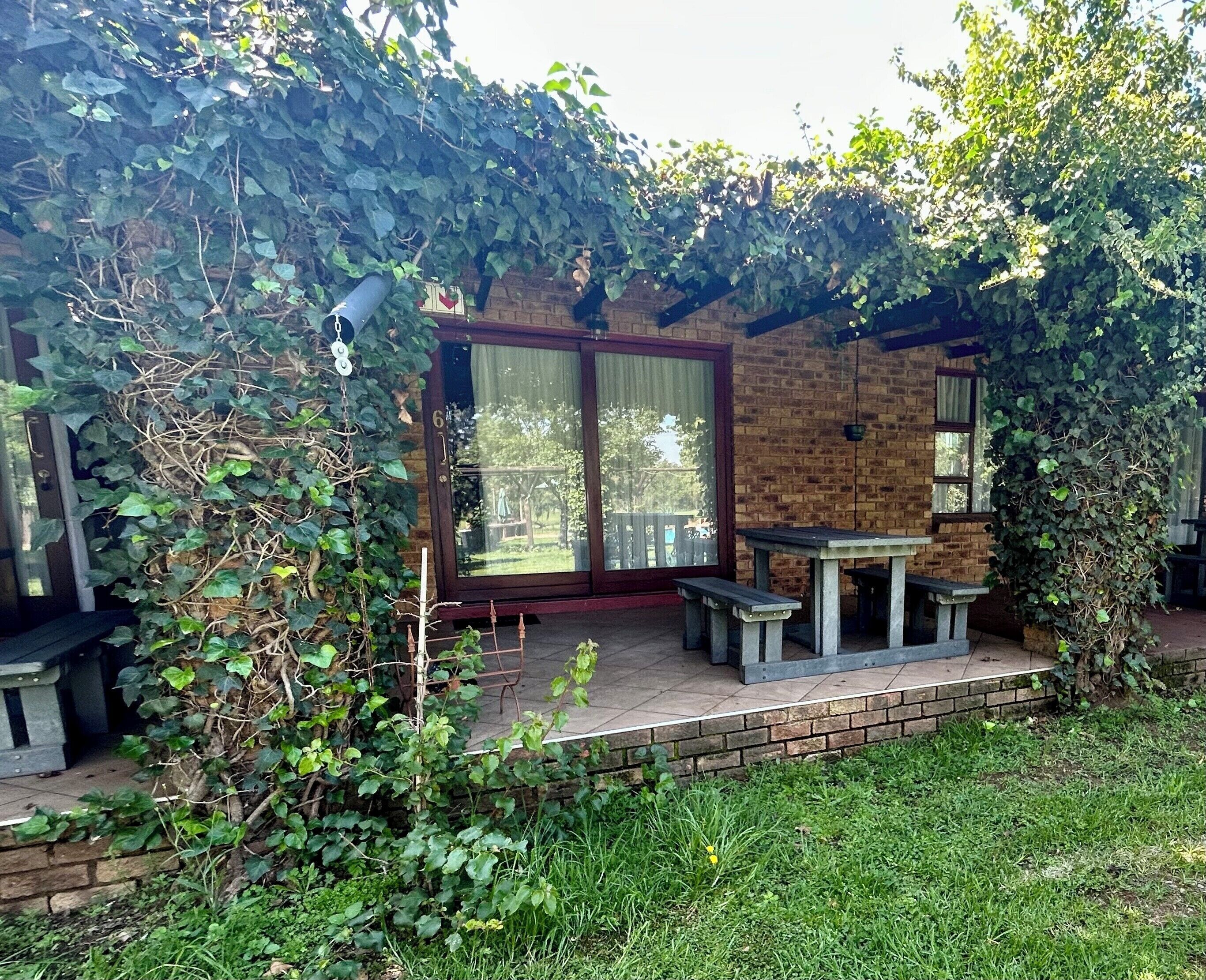 A brick patio with a glass door, surrounded by greenery and a wooden table with benches.