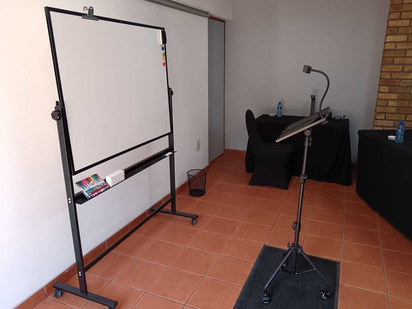 Una habitación con una pizarra, un escritorio con una computadora, una silla y una pequeña mesa con una botella de agua.