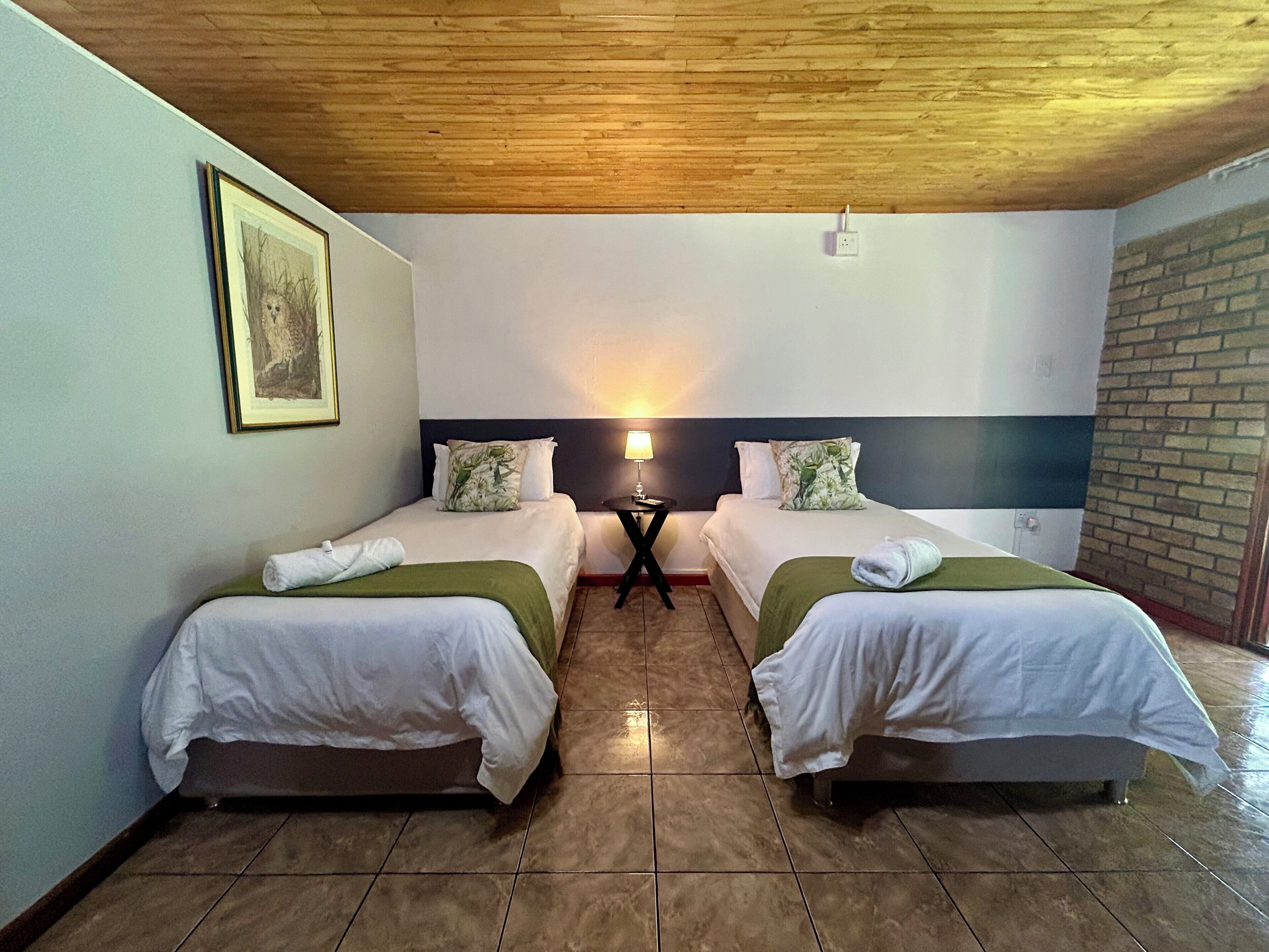 Habitación de hotel con dos camas, una mesita de noche, una lámpara, un cuadro enmarcado y una pared de ladrillo.
