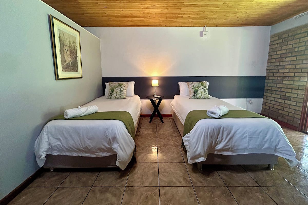 Habitación de hotel con dos camas, una mesita de noche, una lámpara, un cuadro enmarcado y una pared de ladrillo.