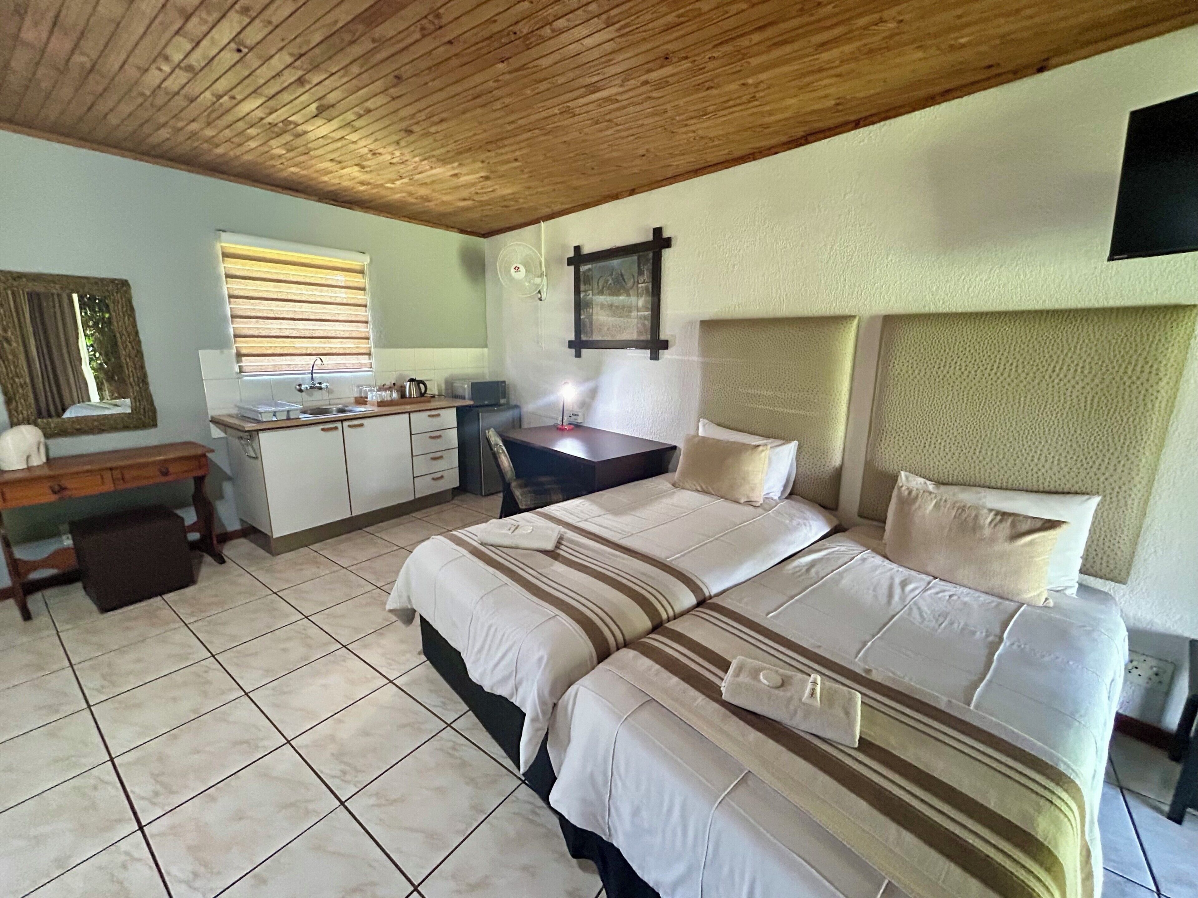 A bedroom with a bed, a desk, a chair, a small kitchenette, and a window with blinds.