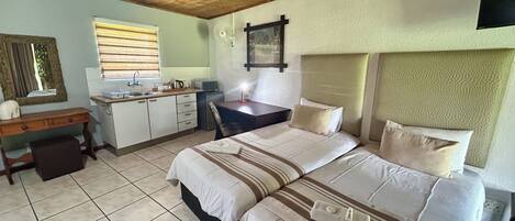 A bedroom with a bed, a desk, a chair, a small kitchenette, and a window with blinds.