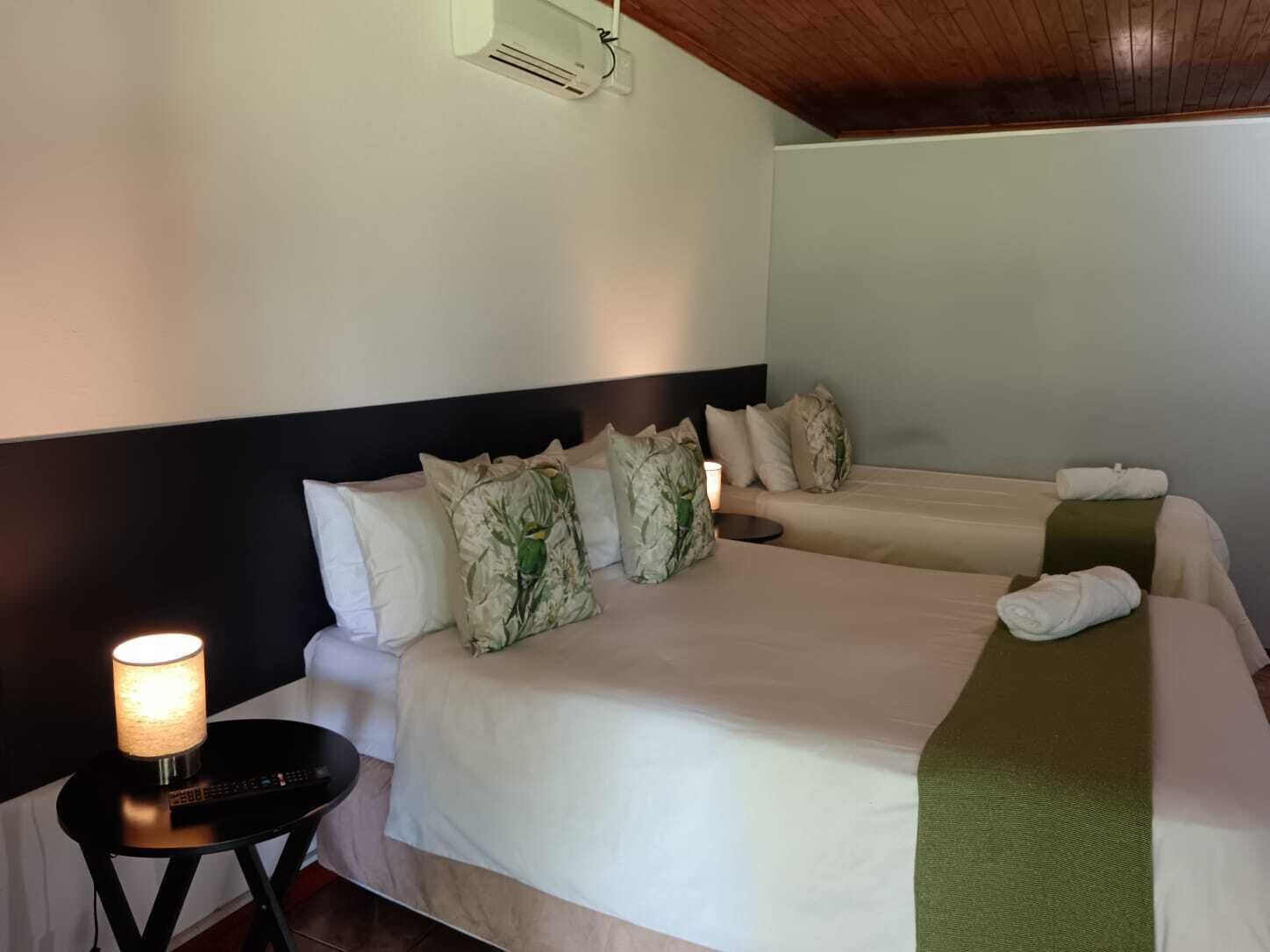 A hotel room with a bed, pillows, a bedside table with a lamp, and an air conditioning unit on the wall.