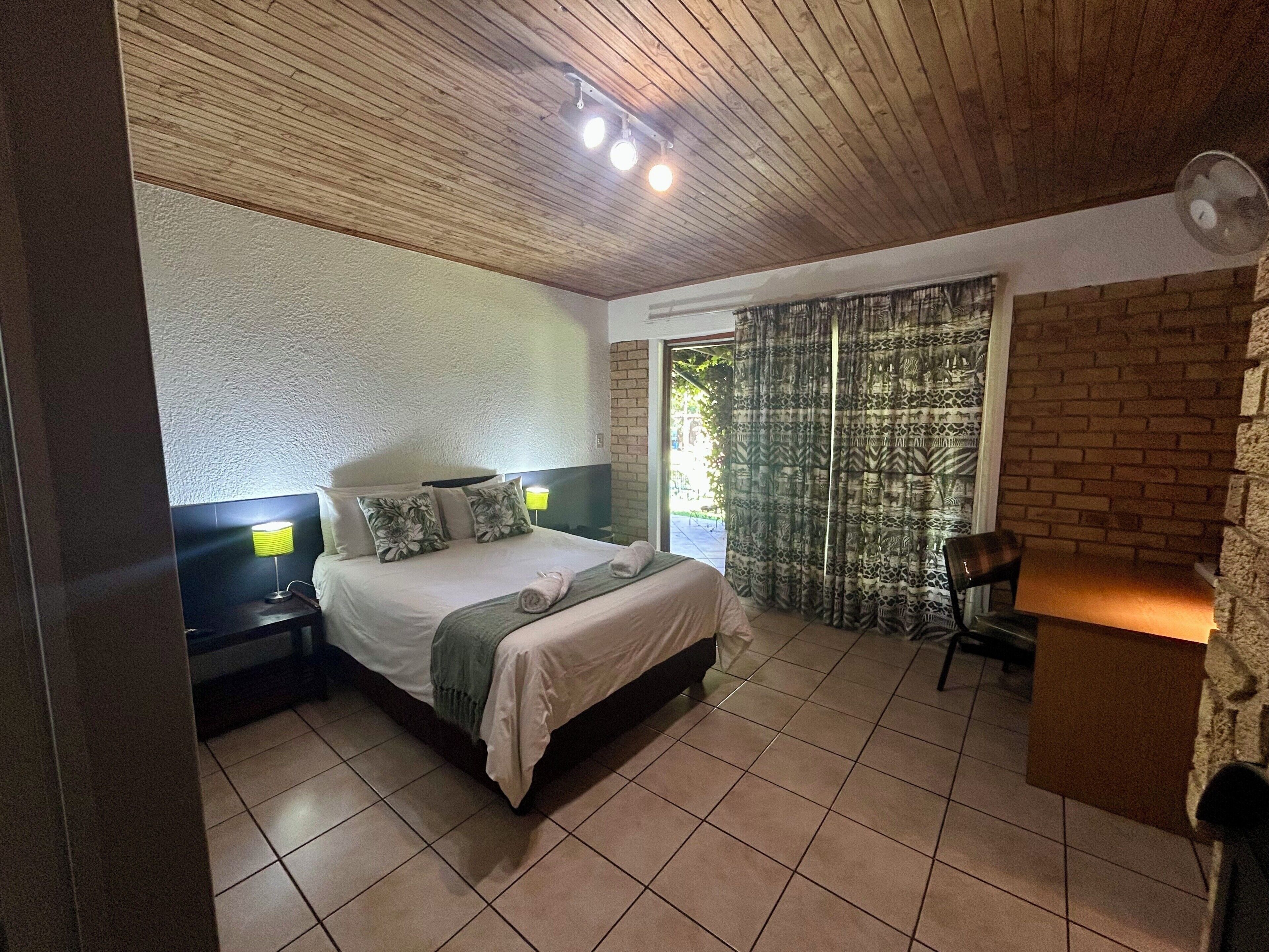 A bedroom with a bed, bedside lamps, a brick wall, a window with curtains, and a fan.