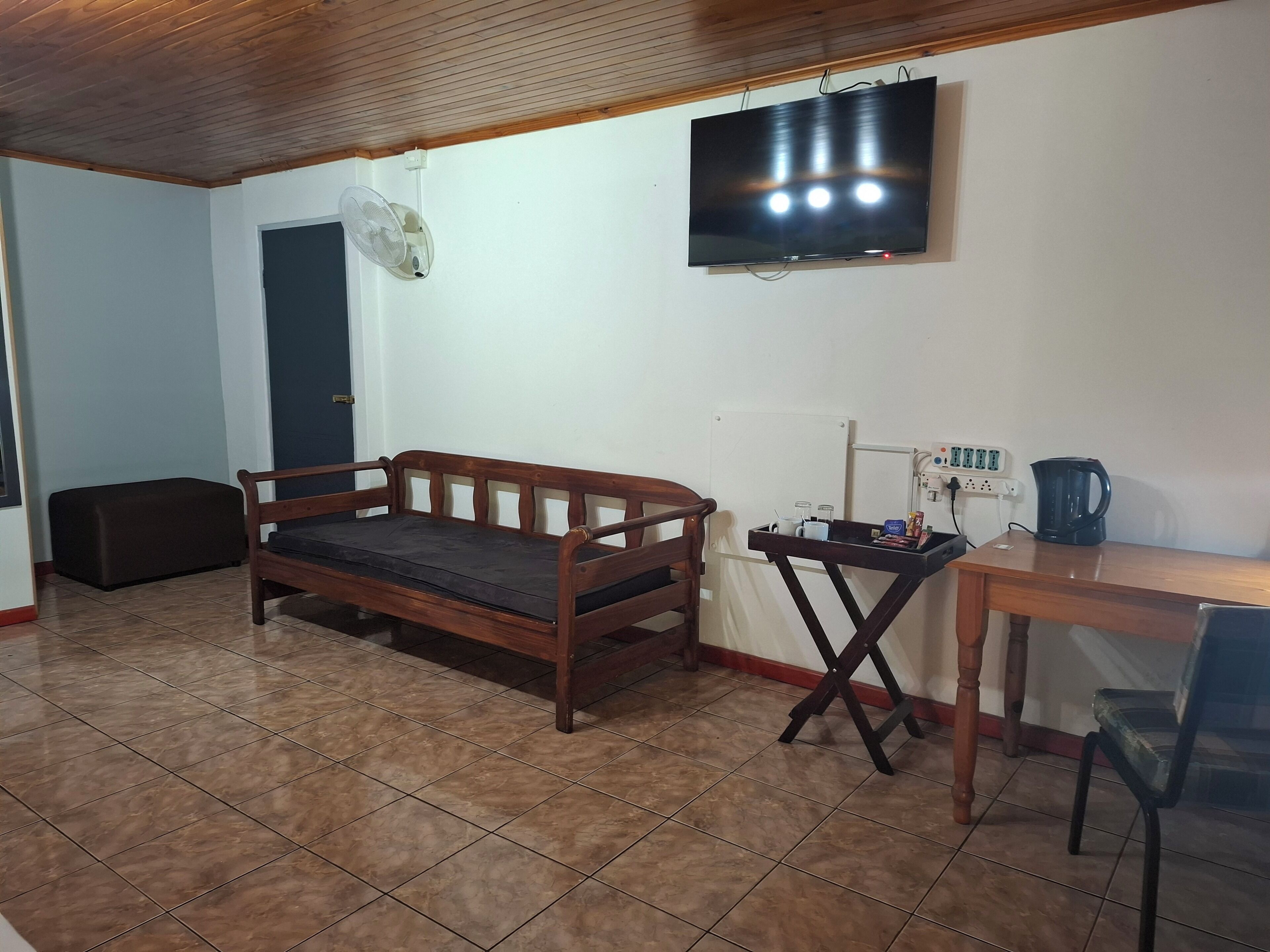 A room with a wooden sofa, a small table with a kettle, a TV, and a fan.