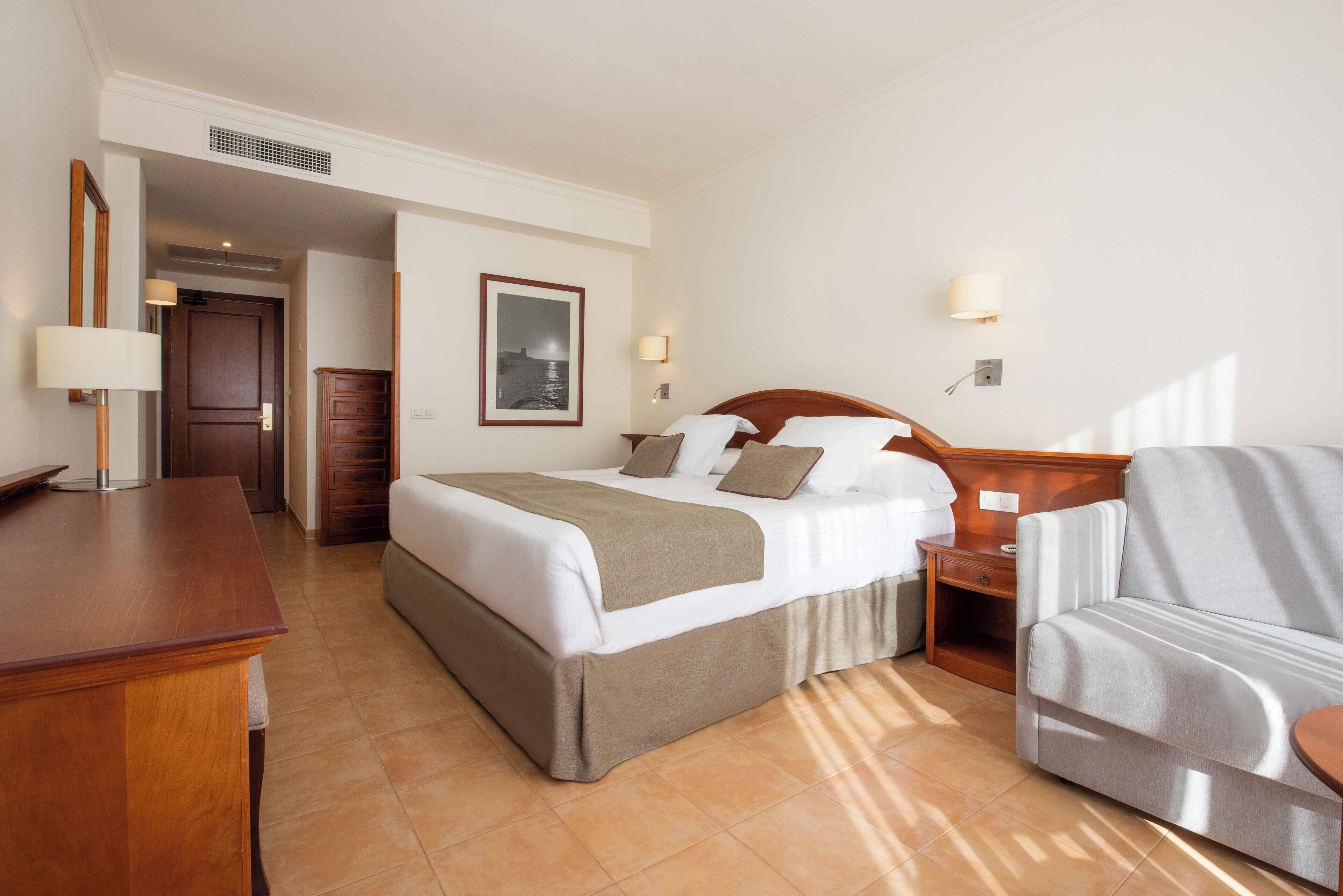 A hotel room with a bed, a desk, a chair, a nightstand, and a wall-mounted lamp.