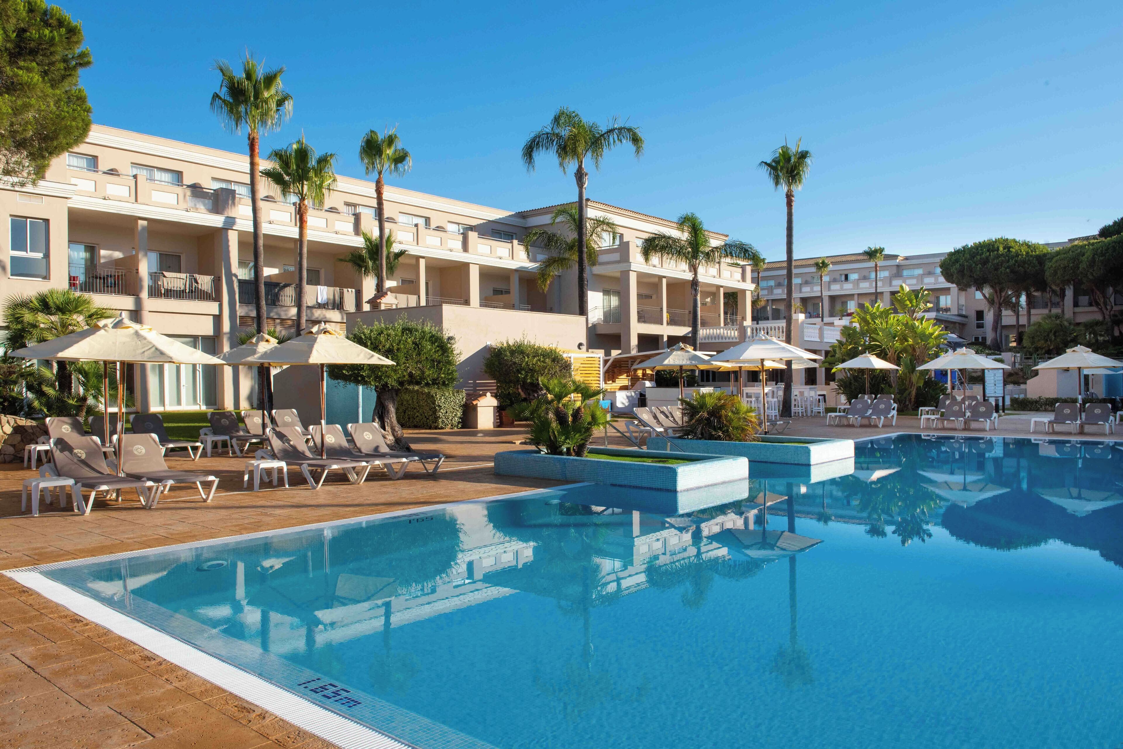 A pool area with lounge chairs and umbrellas, surrounded by a building with balconies and palm trees.