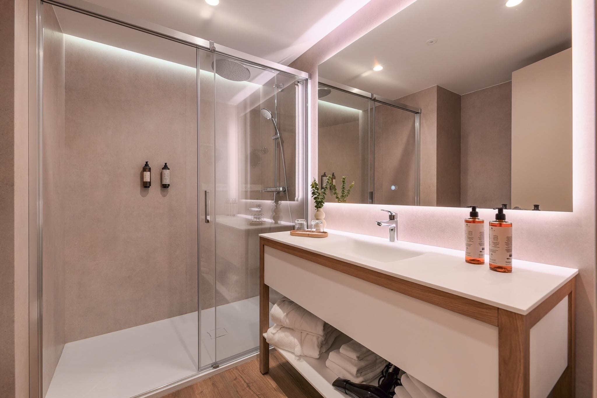 Un baño moderno con cabina de ducha de vidrio, tocador blanco con estructura de madera y un espejo grande.