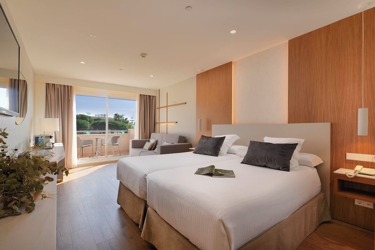 A hotel room with two beds, a balcony with a view, a desk with a chair, and a plant.
