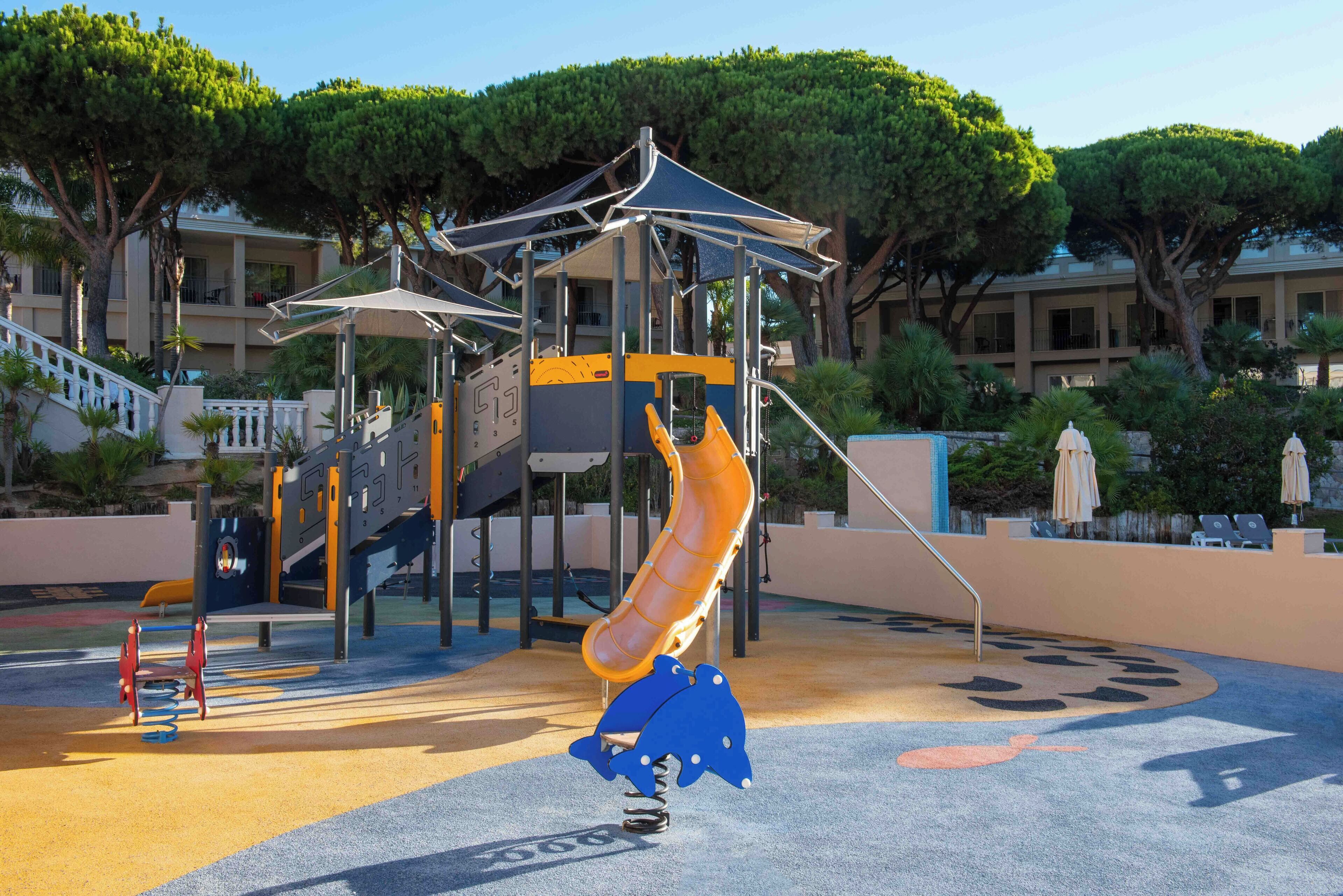 A playground with a slide and climbing structures, surrounded by trees and a building in the background.