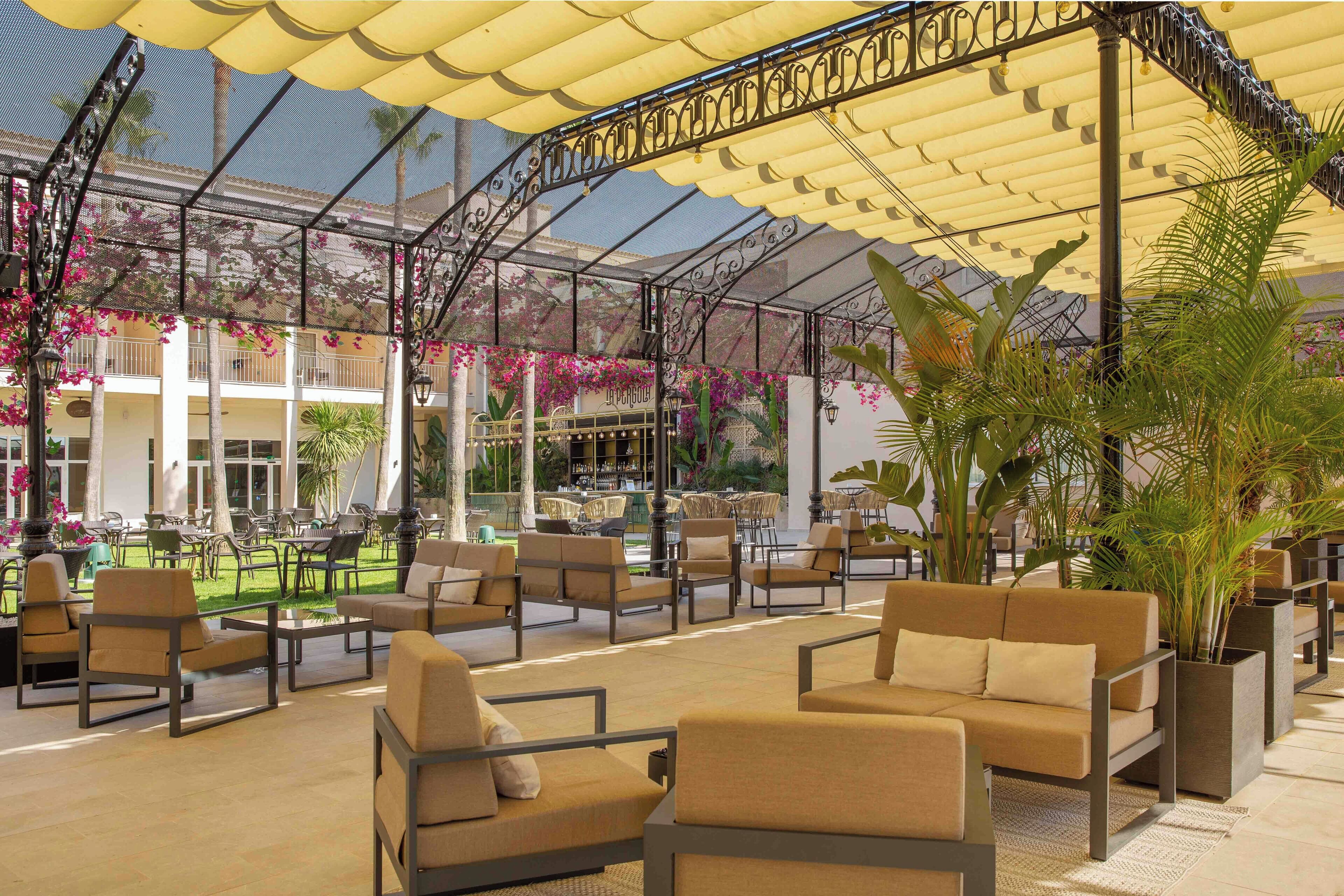 Ein Sitzbereich im Freien mit gepolsterten Stühlen und Tischen, eine überdachte Terrasse mit Metallgeländer und Blumentöpfen.