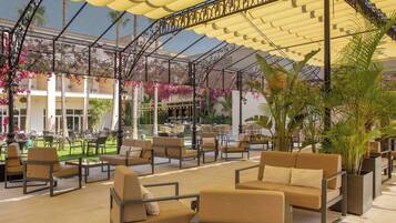 Ein Sitzbereich im Freien mit gepolsterten Stühlen und Tischen, eine überdachte Terrasse mit Metallgeländer und Blumentöpfen.