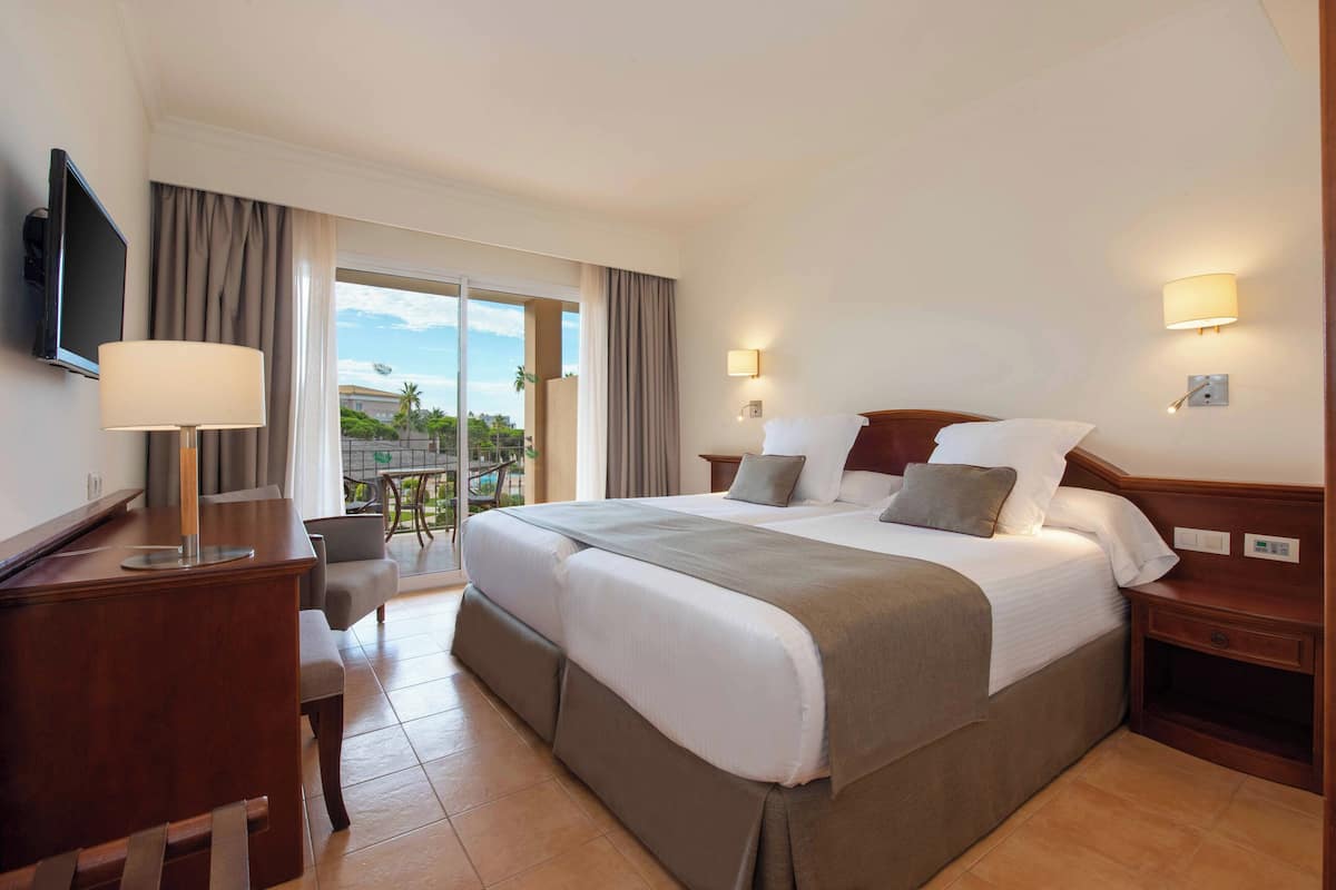 A hotel room with a large bed, a desk, a chair, a TV, and a balcony with a view.