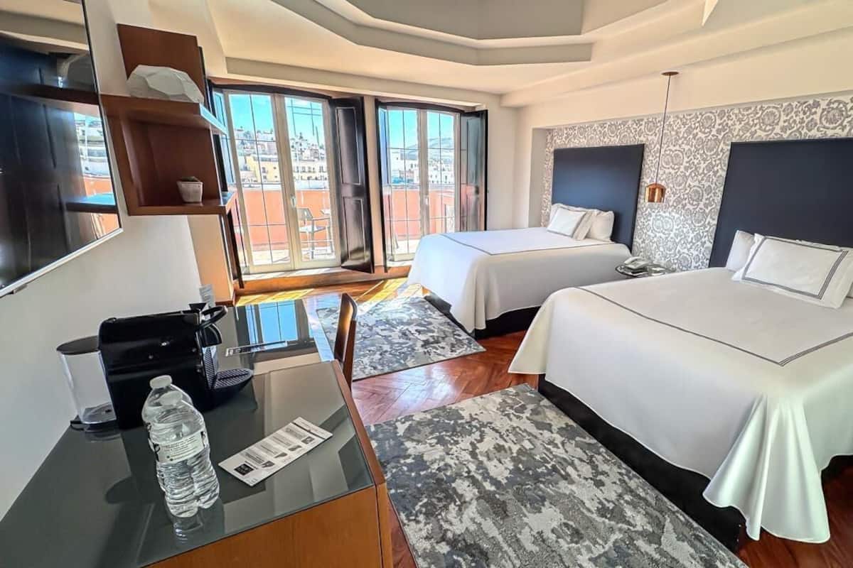 Une chambre d’hôtel avec deux lits, un bureau avec une chaise, une grande fenêtre et un balcon.