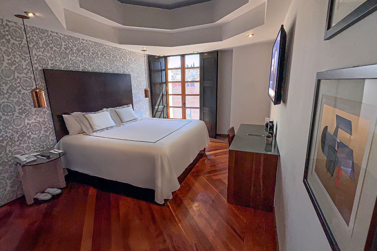 Une chambre à coucher avec un grand lit, une table de chevet, un petit bureau, une fenêtre donnant sur un beau paysage et un papier peint à motifs.