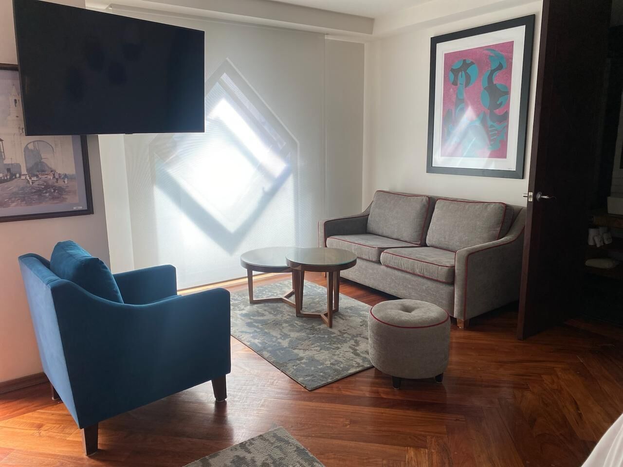 Una sala de estar con un sillón azul, un sofá gris, una mesita de centro redonda y un pequeño otomaní. Se observa un cuadro en la pared y una ventana con persianas.