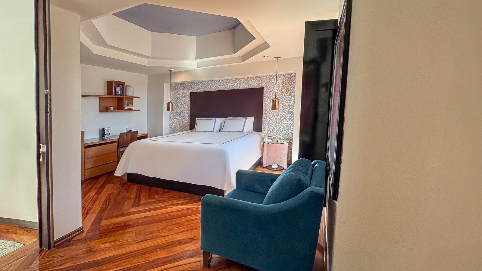 A modern hotel room with a large bed, a blue armchair, a wooden floor, and a patterned wallpaper.
