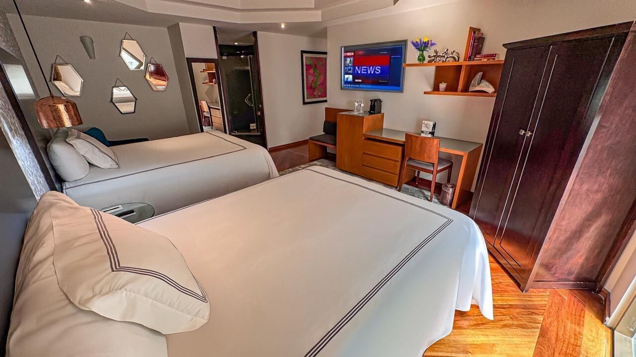 A hotel room with two beds, a desk, a wardrobe, a television, and a wall with geometric mirrors.