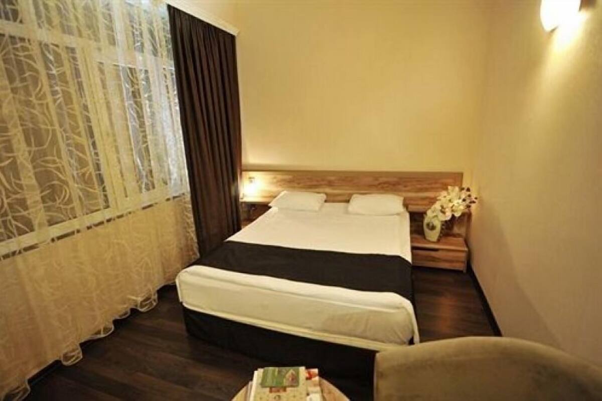 Une chambre d’hôtel avec un lit double, une table de chevet ornée d’un vase de fleurs et une fenêtre avec des rideaux légers.