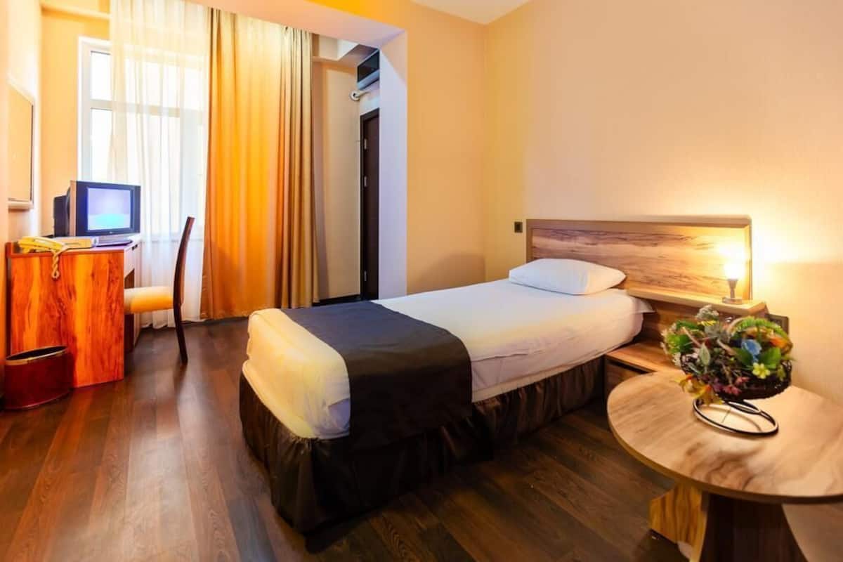 Une chambre d’hôtel avec un lit, un bureau avec un ordinateur, une chaise, une petite table avec une plante et une fenêtre avec des rideaux.