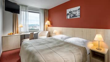 Une chambre d’hôtel avec un lit, deux lampes de chevet, une télévision, une chaise, un bureau avec des fruits et une vue sur la ville.