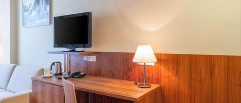 A hotel room with a wooden desk, a television, a lamp, a chair, and a sofa.