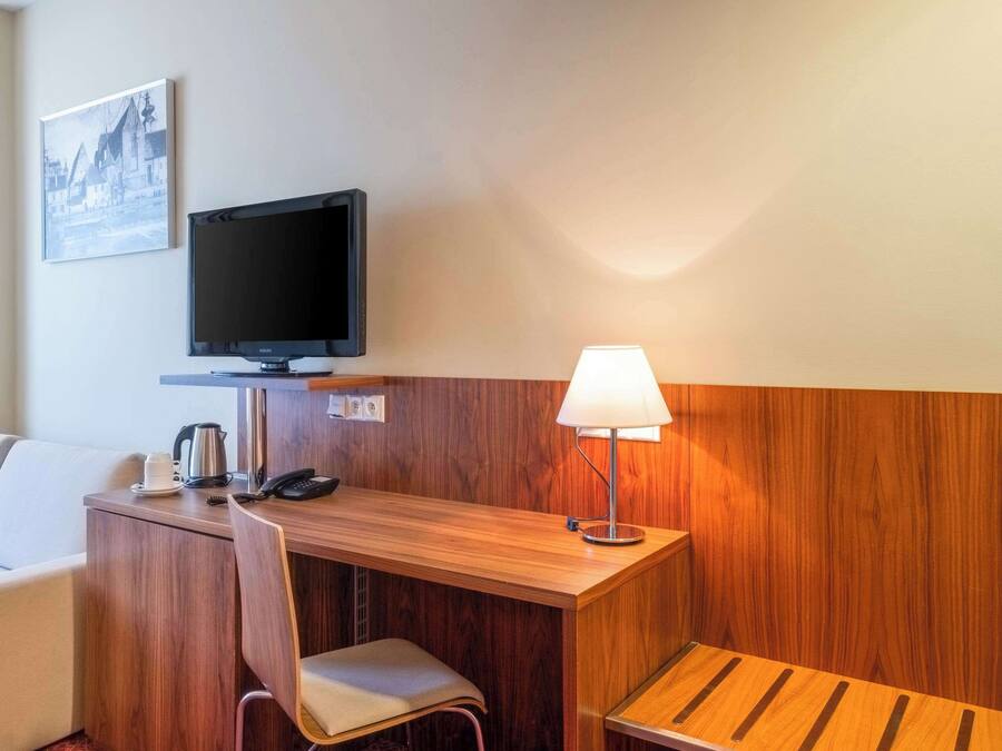 A hotel room with a wooden desk, a television, a lamp, a chair, and a sofa.