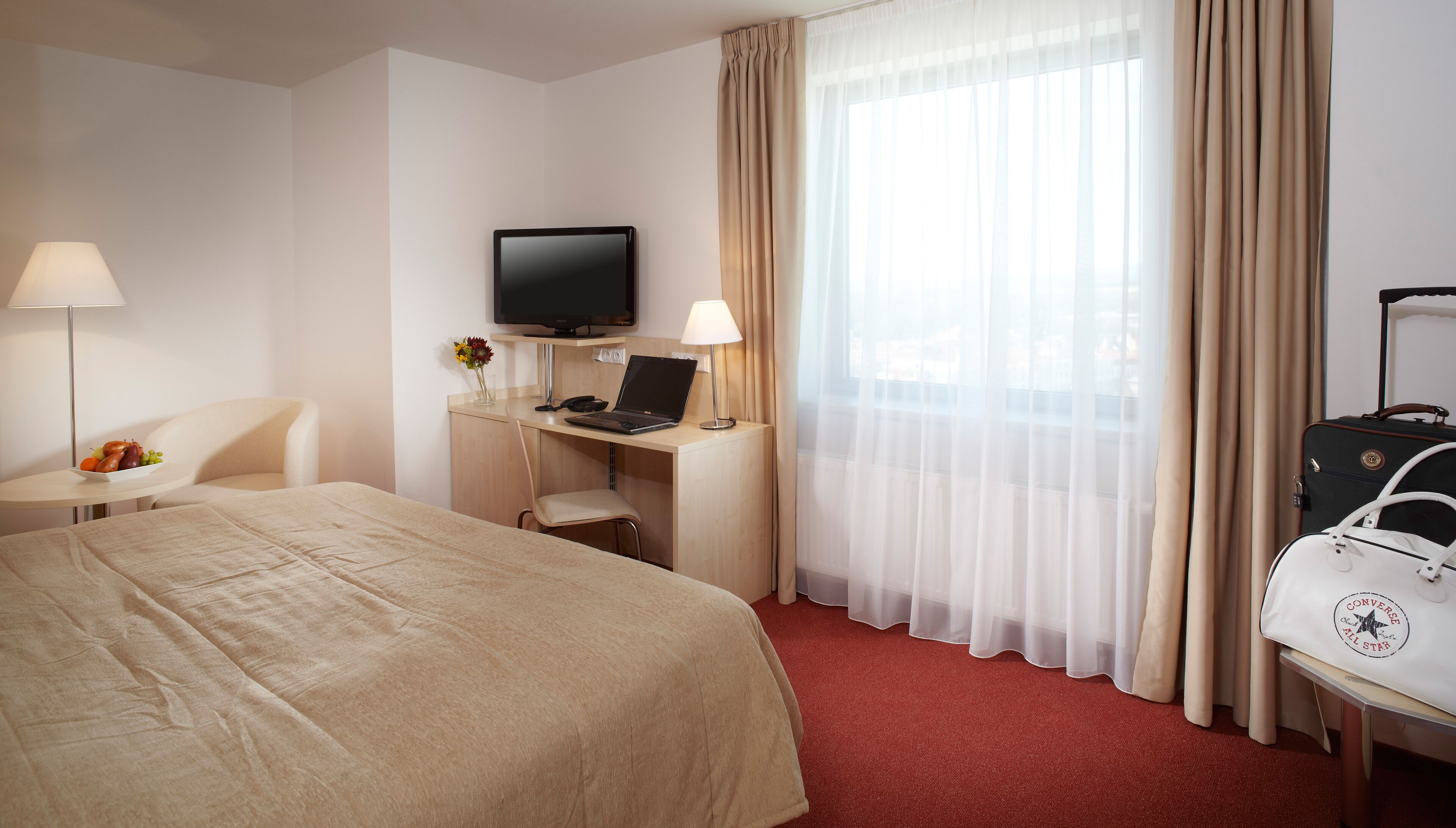 Een hotelkamer met een bed, bureau, laptop, televisie en een koffer.