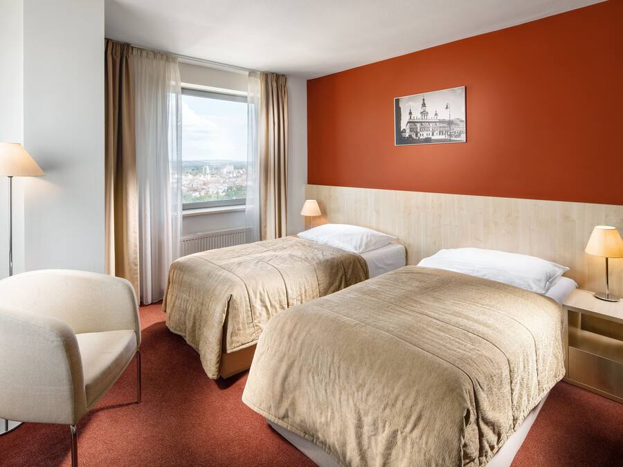 Et hotelværelse med to senge, en stol, et natbord, en lampe og et vindue med gardiner.
