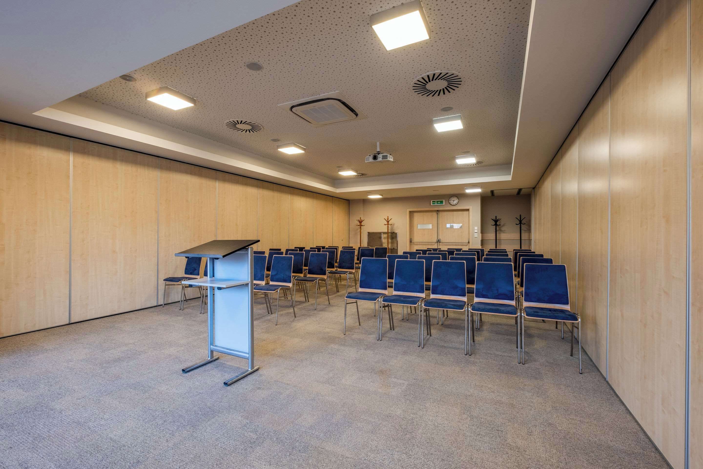 Une salle de conférence avec des rangées de chaises bleues, un pupitre et des luminaires au plafond.