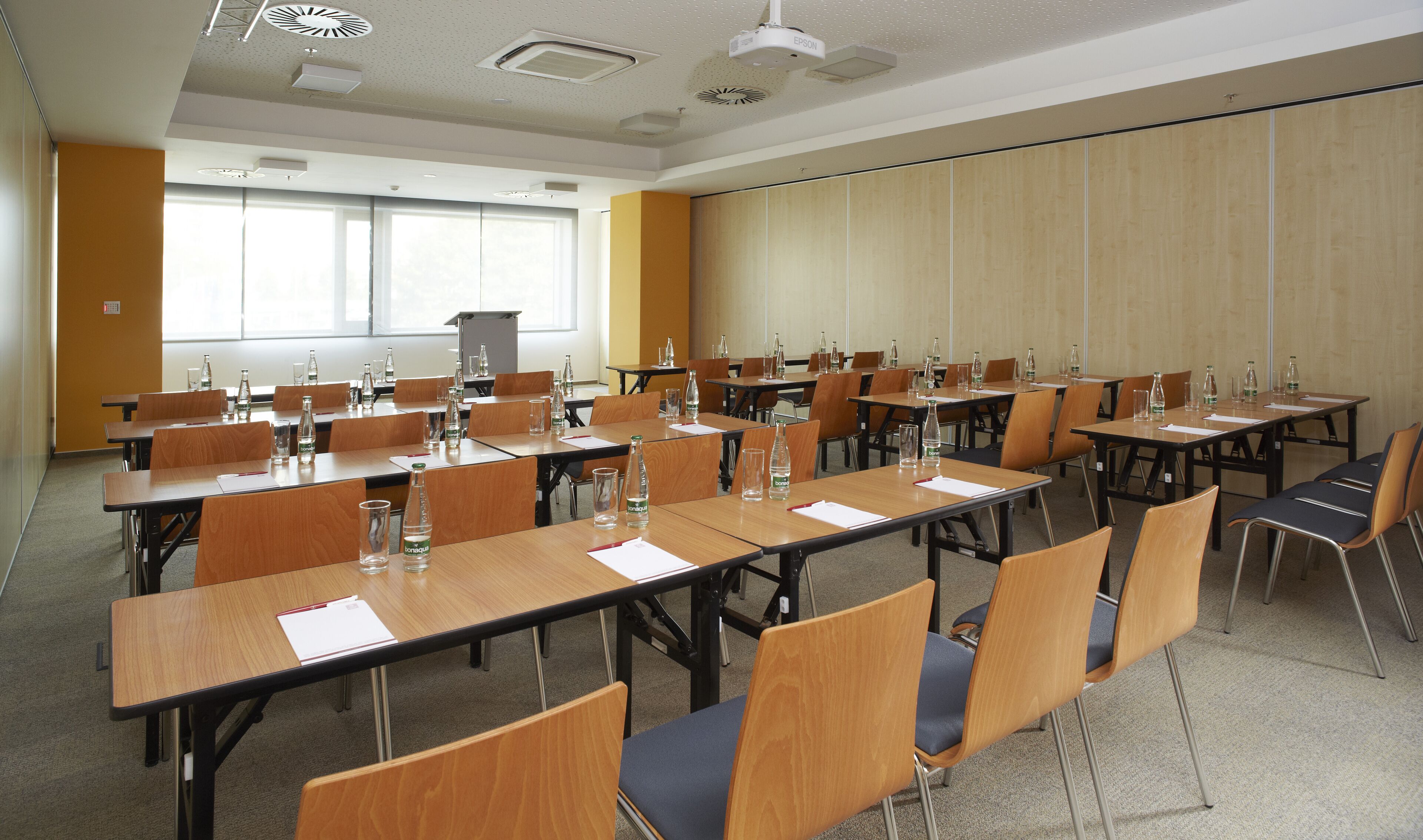 A conference room with multiple tables and chairs, equipped with water bottles and papers, and a projector on the ceiling.