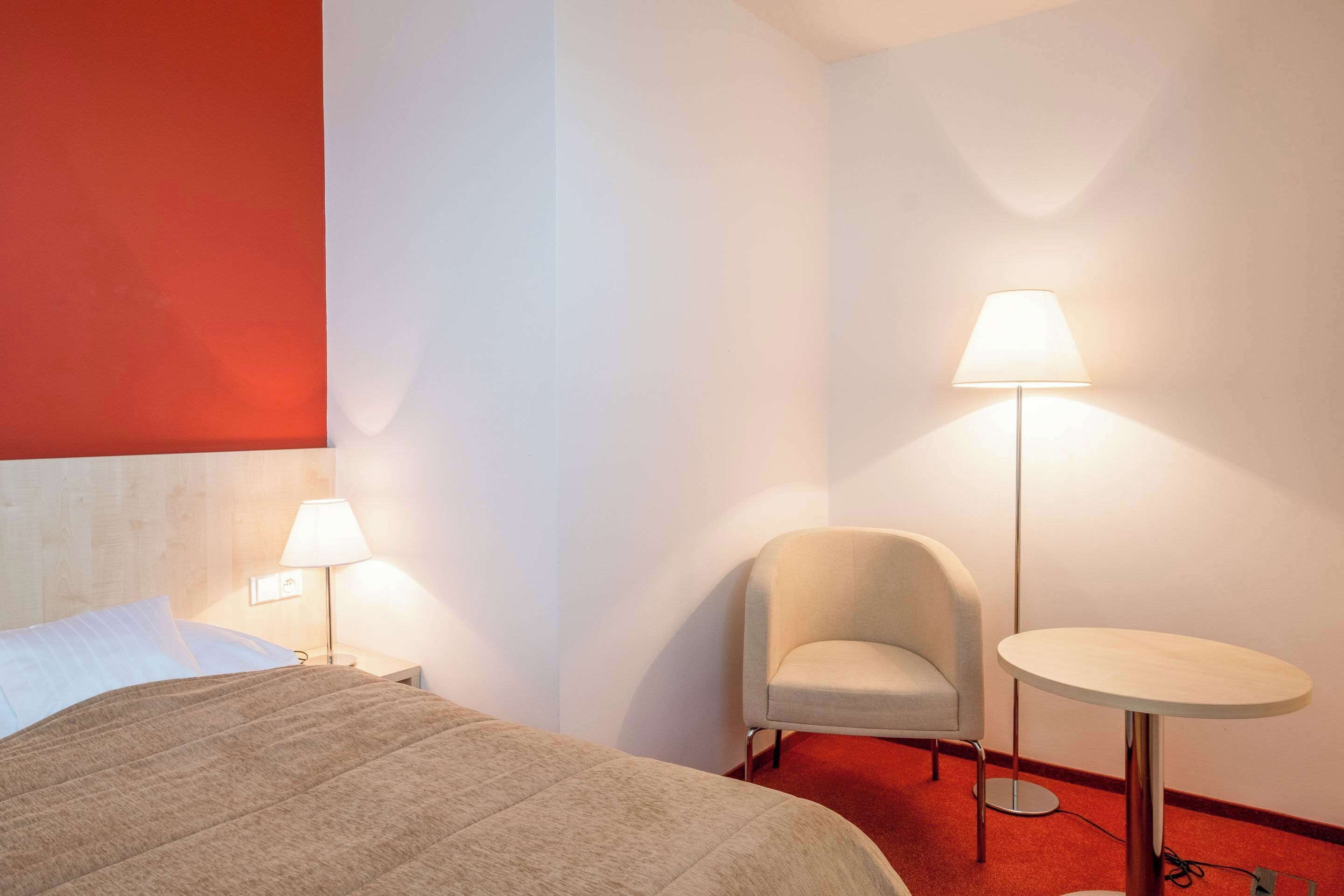 Una habitación de hotel con una cama, una silla, una mesa redonda y dos lámparas de pie.