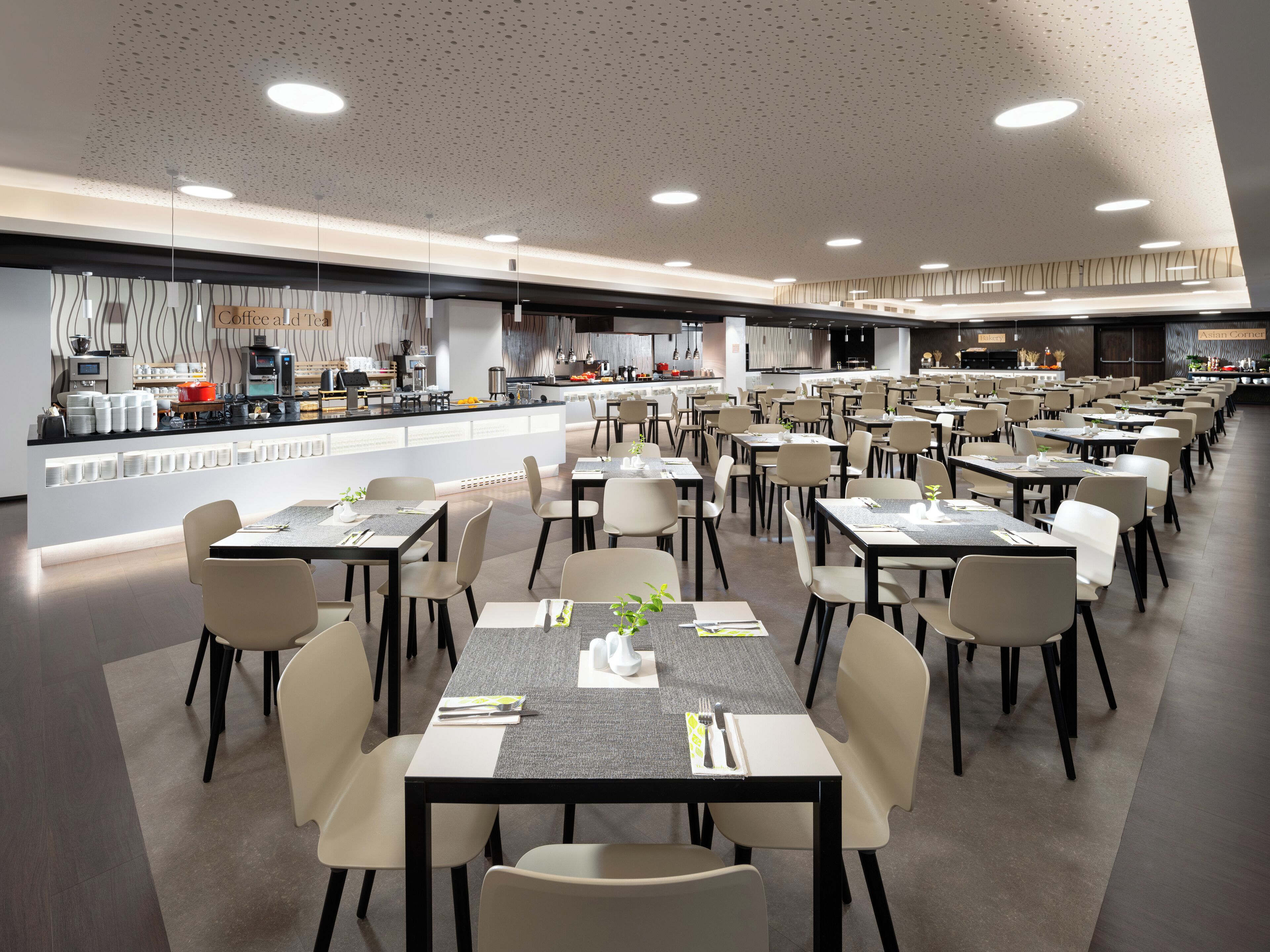 Un comedor moderno con varias mesas y sillas, una cocina al fondo y un techo con iluminación empotrada.