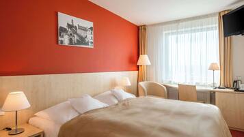 Una habitación de hotel con cama, dos mesitas de noche con lámparas, un escritorio con silla, televisión y ventana con cortinas.