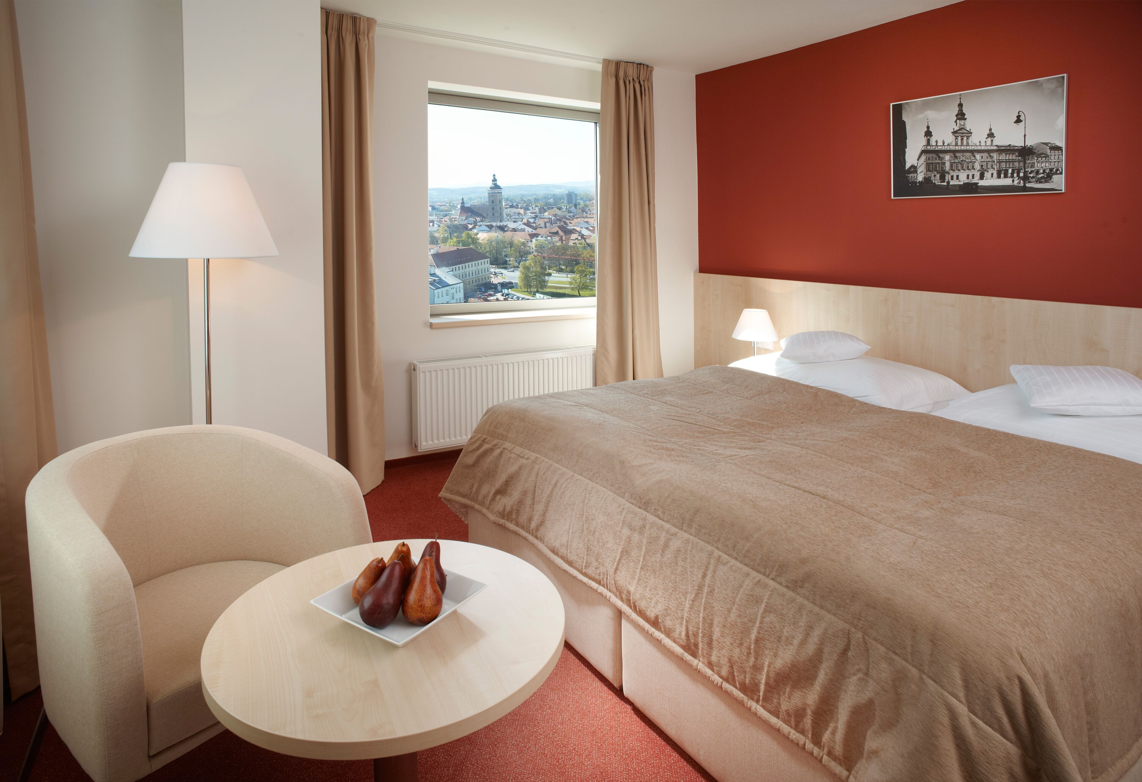 Una habitación de hotel con una cama, una silla, una mesa con fruta, una ventana con vista a un paisaje urbano y un cuadro en la pared.