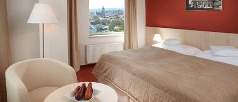 Una habitación de hotel con una cama, una silla, una mesa con fruta, una ventana con vista a un paisaje urbano y un cuadro en la pared.