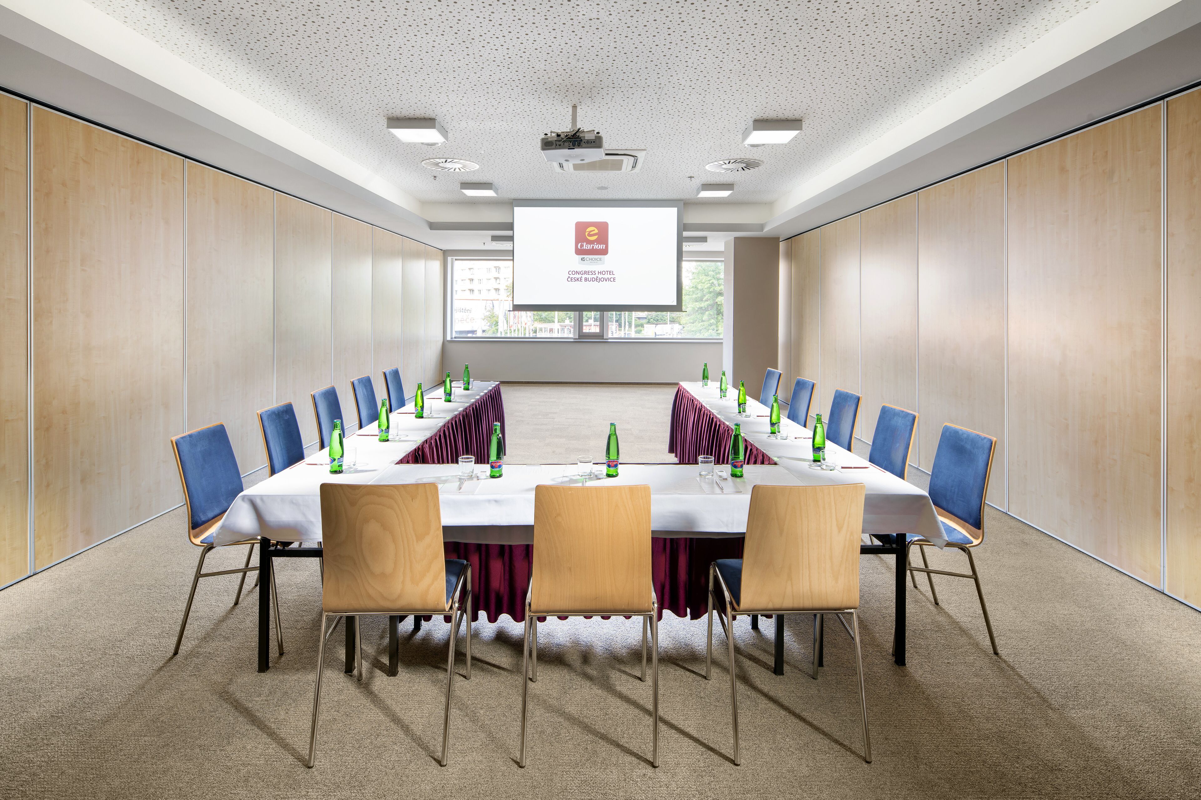 A conference room with a long table, blue and wooden chairs, and a projector screen at the front.