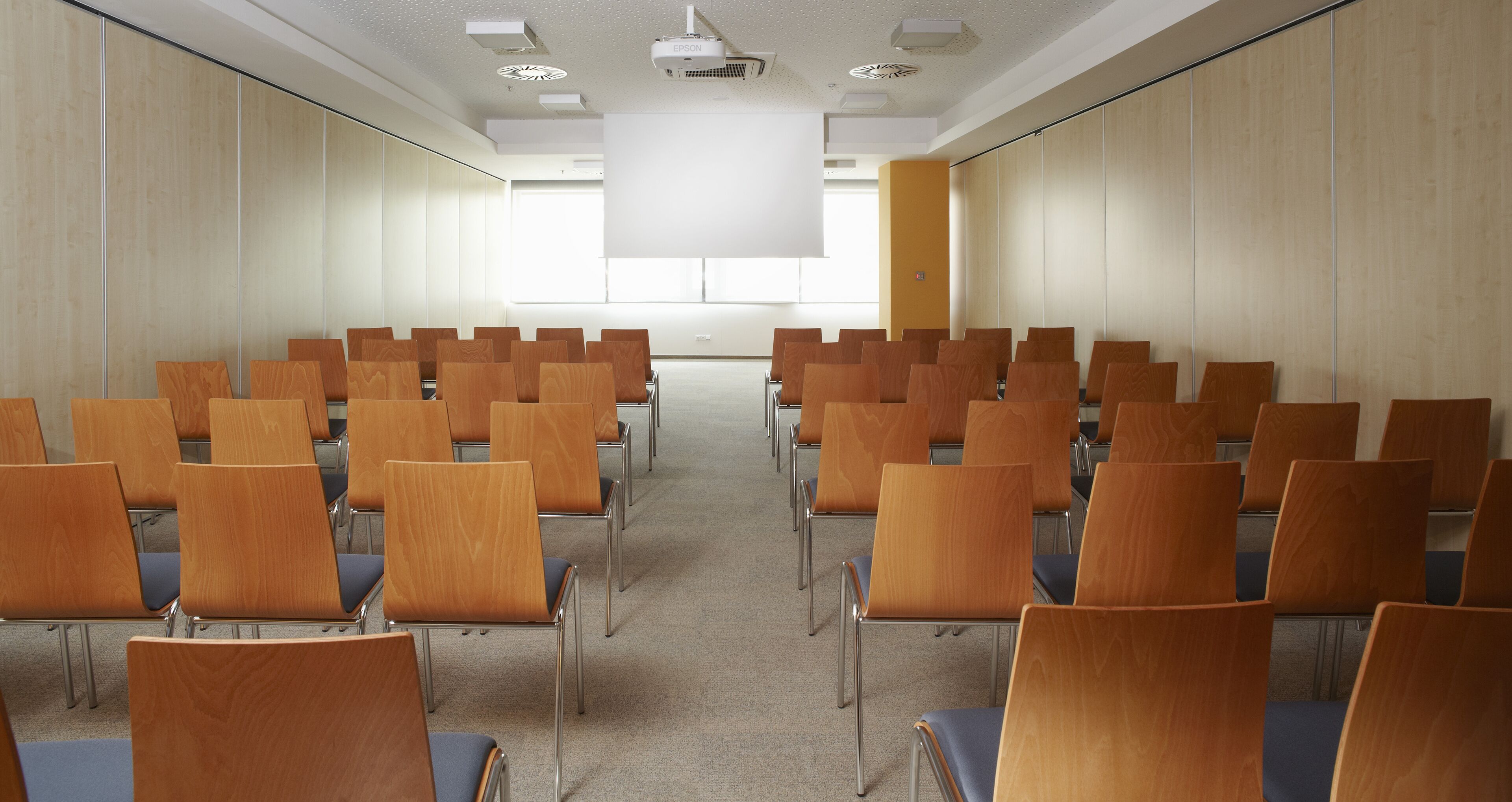 A conference room with rows of wooden chairs, a projector screen, and a large window.