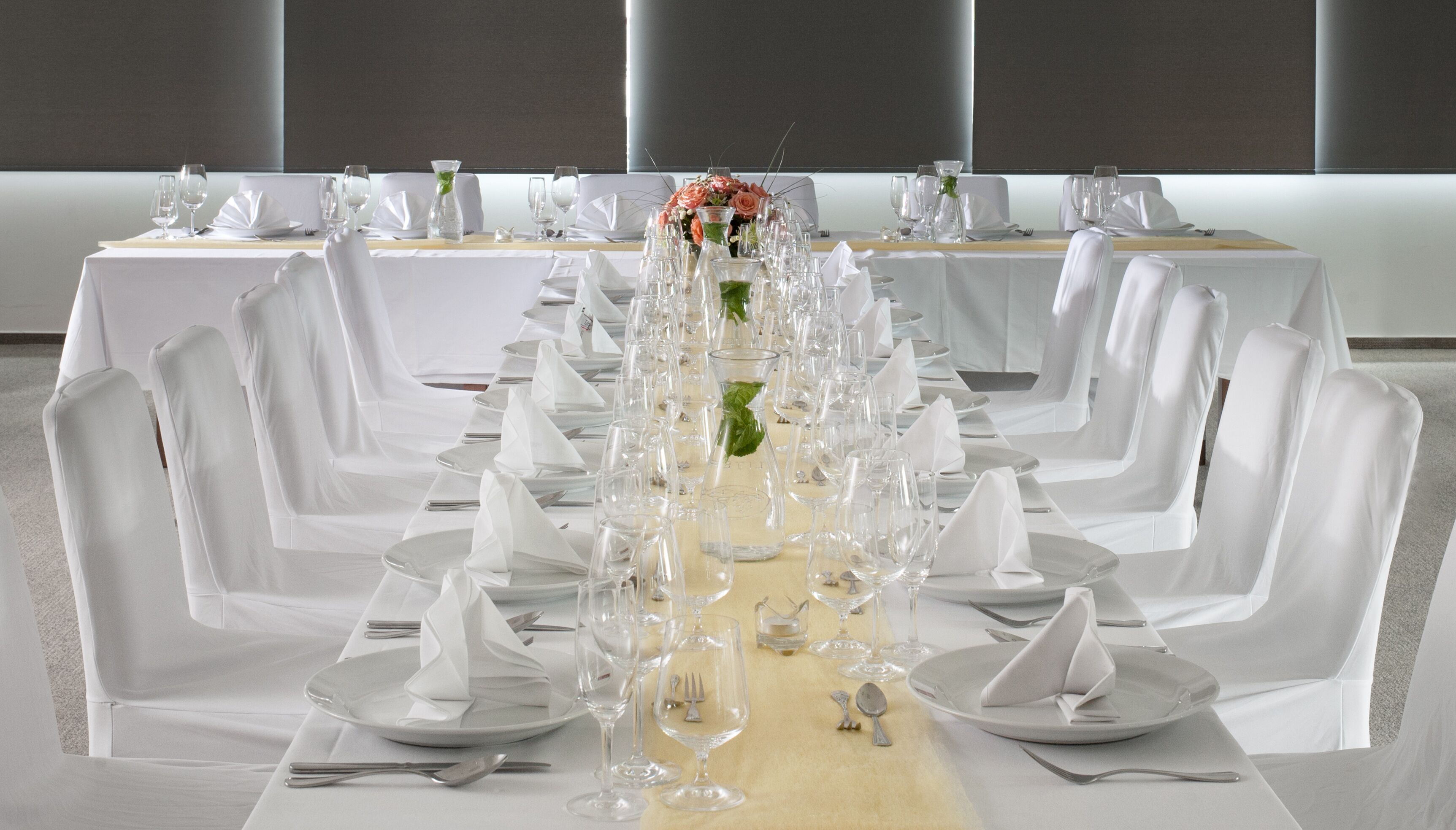 Una larga mesa de banquete dispuesta con manteles blancos, sillas y cristalería, con un centro de mesa floral.