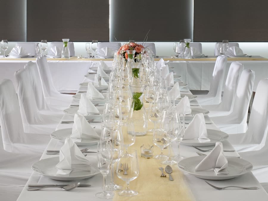 Una larga mesa de banquete dispuesta con manteles blancos, sillas y cristalería, con un centro de mesa floral.